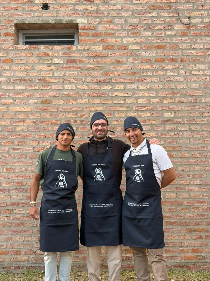 Tres integrantes del equipo, con delantal y gorro de Madre del Pan, sonríen abrazados frente a una pared de ladrillo